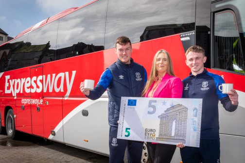 Three people are standing in front of a Bus Éireann Expressway red coach holding a prop of a fiver and cups